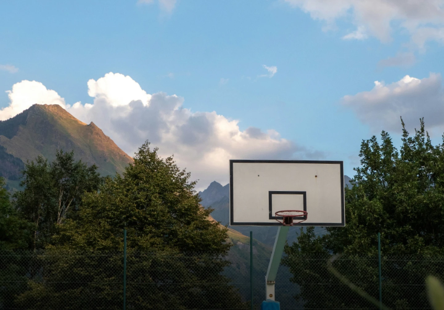 Basketball hoop against backdrop of trees, mountains, and clouds in sky.